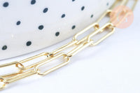 Close-up of Collier Chaine laiton doré clair maille rectangle, featuring a shiny gold chain on a white surface with a polka-dotted ceramic object in the background.
